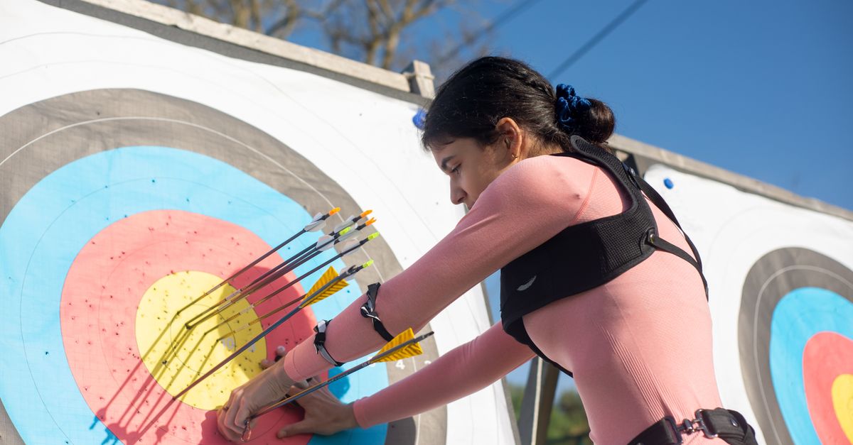 A female archer adjusts arrows on a target during a sunny day, showcasing precise archery skills.